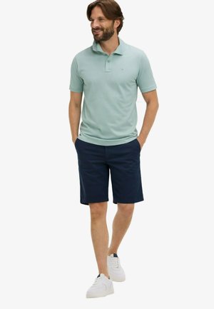 Homme marchant les mains dans les poches, portant un polo vert clair, un short bleu marine et des baskets blanches, souriant et regardant sur le côté.