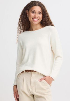 Femme aux cheveux bouclés portant un pull blanc à manches longues et un pantalon beige clair avec une ceinture tressée marron, souriante avec une main dans la poche.