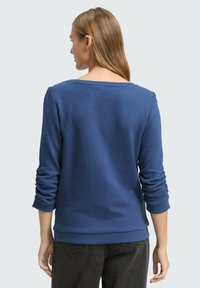 Blauer, langärmeliger Pullover mit weitem Halsausschnitt und gerippten Bündchen. Weicher Stoff und figurbetonter Saum, von hinten gesehen.