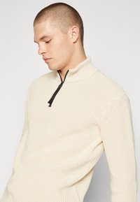 Jersey de color crema con cuello alto y cierre de media cremallera, que presenta un patrón texturizado y puños acanalados.