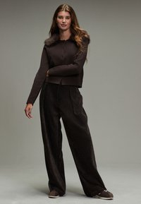 Brązowy krótki kardigan z kołnierzem z sztucznego futra, w zestawie z luźnymi brązowymi spodniami sztruksowymi o teksturowanej powierzchni.