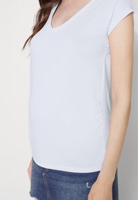 T-shirt branca de decote em V, com mangas curtas e detalhe franzido na lateral, combinada com calções de ganga. Tecido suave, corte casual.