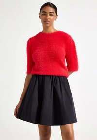 Pull rouge duveteux à manches courtes avec un col rond, associé à une jupe noire évasée. La texture du pull est moelleuse et douce.