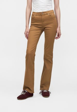 Femme portant un pantalon flare marron ajusté, un haut blanc froncé et des chaussures à lacets rouge foncé, debout devant un fond clair uni.