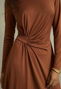 Abito lungo marrone con maniche lunghe e dettaglio a nodo attorcigliato in vita. Tessuto morbido con una texture liscia, silhouette fluida e design minimale.