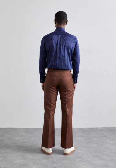 Chemise bleue rayée à boutons avec un dos ajusté, associée à un pantalon marron légèrement évasé aux poignets, complétée par des baskets blanches.