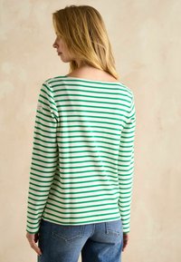 zaļš un balts svītrots garām piedurknēm krekls ar plašu izgriezumu. Audums izskatās mīksts; uz kreisā pleca ir logo uzlīme. Apvienots ar ziliem džinsiem.