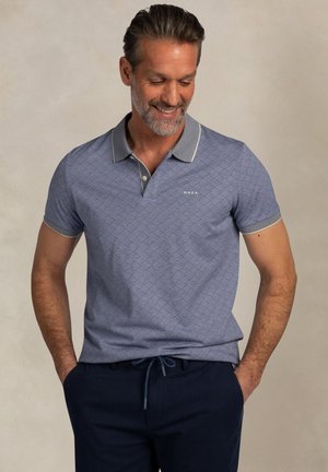 Man glimlachend en omlaag kijkend, gekleed in een blauwe poloshirt met patroon en donkerblauwe broek, met handen in de zakken tegen een effen achtergrond.