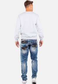 Sweatshirt gris avec des poignets côtelés, associé à un jean bleu déchiré présentant de multiples poches, des textures variées et des détails de couture contrastants.
