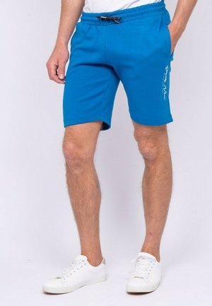 Homme portant un short bleu vif avec cordon de serrage et poches latérales, ainsi que des baskets blanches, debout devant un fond uni clair.