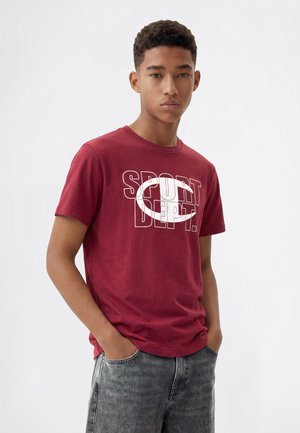 Jeune homme aux cheveux courts et bouclés, portant un t-shirt rouge avec le texte "SPORT DEPT." et un jean gris, debout les mains dans les poches.