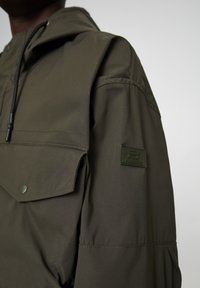Olijfgroene hoodie met een voorzak, drukknoopsluiting en een textuur logo patch op de bovenarm. Gladde stoffen afwerking.
