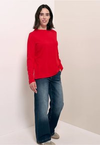 Rotes langärmeliges Oberteil in Slim Fit, aus leichtem Stoff, kombiniert mit blauen weit geschnittenen Jeans, die ein entspanntes Design und subtile Ausbleichungen aufweisen.