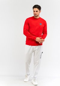 Felpa rossa concollo rotondo e patch con logo, abbinata a pantaloni della tuta bianchi con testo e dettagli neri, completata da sneakers bianche.