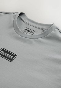 Szürke pamut póló kerek nyakkivágással, az elején fekete téglalap alakú „oasis” feliratú logóval. Simább felület, hétköznapi kialakítás.