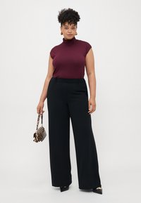 Mujer vistiendo un suéter sin mangas de color burdeos, pantalones anchos negros, zapatos de punta negros, aretes dorados, sosteniendo un pequeño bolso de mano de estampado de leopardo.