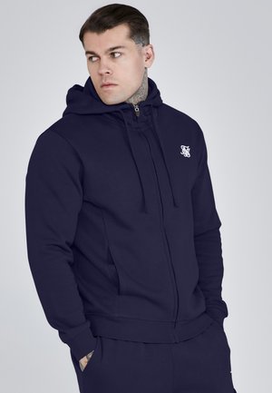 Homme avec des piercings aux oreilles et au nez, portant un sweat à capuche bleu marine zippé avec un petit logo blanc sur la poitrine, regardant vers sa gauche.