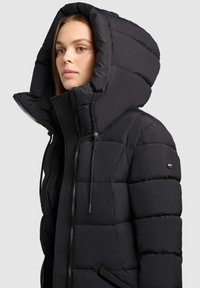 Chaqueta de plumas negra con una gran capucha, cierre frontal con cremallera y cordones elásticos. Presenta un texturizado acolchado y un logo en la manga.