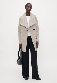 Beige wolmix jas met een brede kraag, open voorkant en twee zwarte knopen. Geschreven over een witte top en gecombineerd met zwarte wijde pantalons en accessoires.