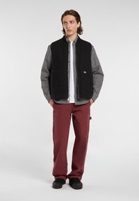 Gilet nero con zip sopra una camicia a quadri, abbinato a pantaloni bordeaux. Il gilet presenta due tasche frontali e un'etichetta con il logo. Tessuto texturizzato.