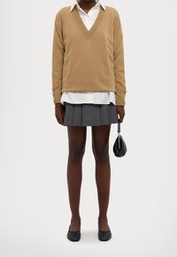 Pull beige à col en V sur une chemise blanche, mini-jupe plissée grise, ballerines noires, tenant un petit sac noir. Texture lisse, design décontracté.