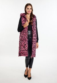 Vesta cu imprimeu de leopard roz, umplută cu puf, cu glugă, având fermoar în față și buzunare laterale, purtată peste un top negru și pantaloni din piele.