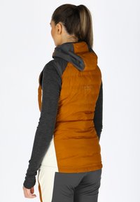 Gilet imbottito arancione con cappuccio grigio e dettagli sulle spalle. Tessuto a trama evidenziata con design a righe verticali. Il modello indossa una maglietta a maniche lunghe grigia.