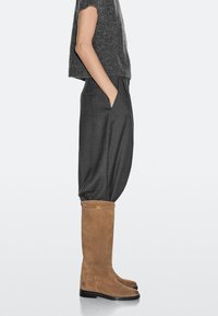 Stivali in camoscio marrone fino al ginocchio con punta arrotondata, suola piatta nera e dettagli in oro. Indossati con pantaloni grigi larghi e un maglione lavorato a maglia.