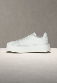 Baskets basses blanches à plateforme en cuir texturé, fermeture à lacets et bout arrondi, présentées sur une surface ronde avec un fond neutre.