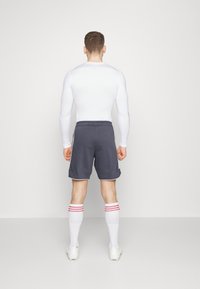 Witte langslobige sporttop, grijze shorts met zijaccenten en kniehoge witte sokken met rode strepen; neutrale achtergrond.