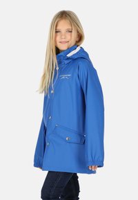 Jeune fille aux longs cheveux blonds portant une veste de pluie à capuche bleue et un jean foncé, debout de profil et souriant légèrement.