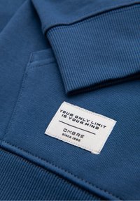 Manche et poche de sweat bleu avec une étiquette blanche indiquant "Your only limit is your mind" et le nom de la marque Ombre depuis 1985.