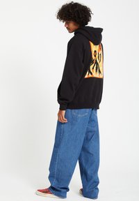 Zwarte hoodie met een levendig, abstract grafisch ontwerp op de achterkant. Gecombineerd met oversized blauwe denim jeans en meerkleurige sneakers.