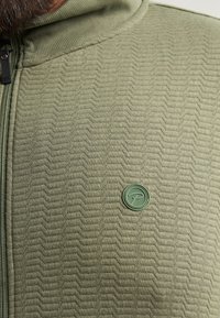 Chaqueta verde oliva con cremallera que presenta un patrón texturizado ondulado, una insignia de logo redonda y detalles acanalados en el cuello y la línea de la cremallera.