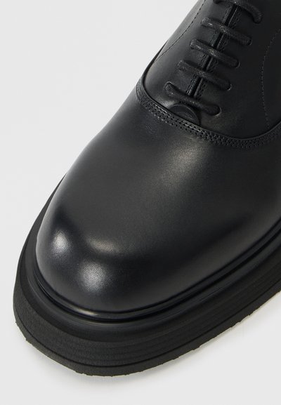 Black leather shoe with a polished surface, rounded toe, and lace-up design. Features a thick rubber sole with horizontal grooves.