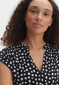 Zwarte blouse met een geometrisch wit vierkant patroon, korte mouwen en een v-hals. Lichtgewicht stof, gladde textuur en een ontspannen pasvorm.