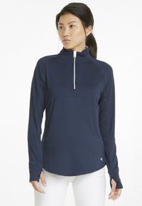 Donna che indossa un top zip-up a maniche lunghe blu navy con fori per le dita e pantaloni bianchi, in piedi davanti a uno sfondo bianco semplice.