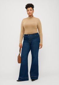 Beige langermet topp parret med blå vide bukser. Modellen holder en brun handlenett. Glatt stoff og strukturert form, avslappet stil.