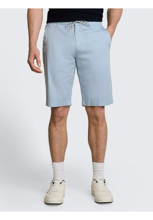 Mann trägt hellblaue knielange Shorts mit Seitentaschen, weiße Socken und weiße Sneakers und steht vor einem einfarbigen Hintergrund.