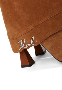 Stivale alla caviglia in suede marrone con un originale tacco triangolare e un accento del logo "Karl" in argento vicino alla base. Texture liscia e design moderno.