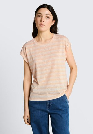 Frau mit langen dunklen Haaren, die ein kurzärmeliges pfirsichfarben gestreiftes T-Shirt und blaue Jeans trägt, steht mit einer Hand in der Tasche.