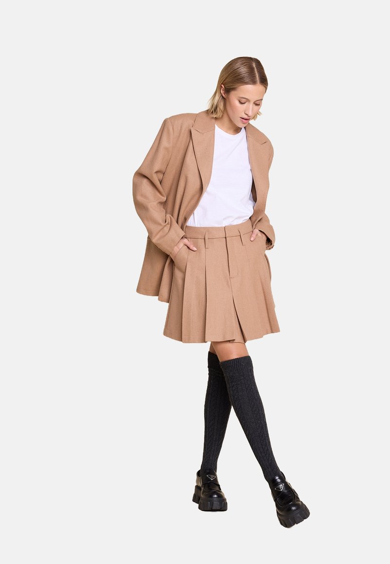 Beige ullblazer over en hvit t-skjorte, kombinert med en plissert beige skjørt, grå knelange sokker, og kraftige svarte sko.