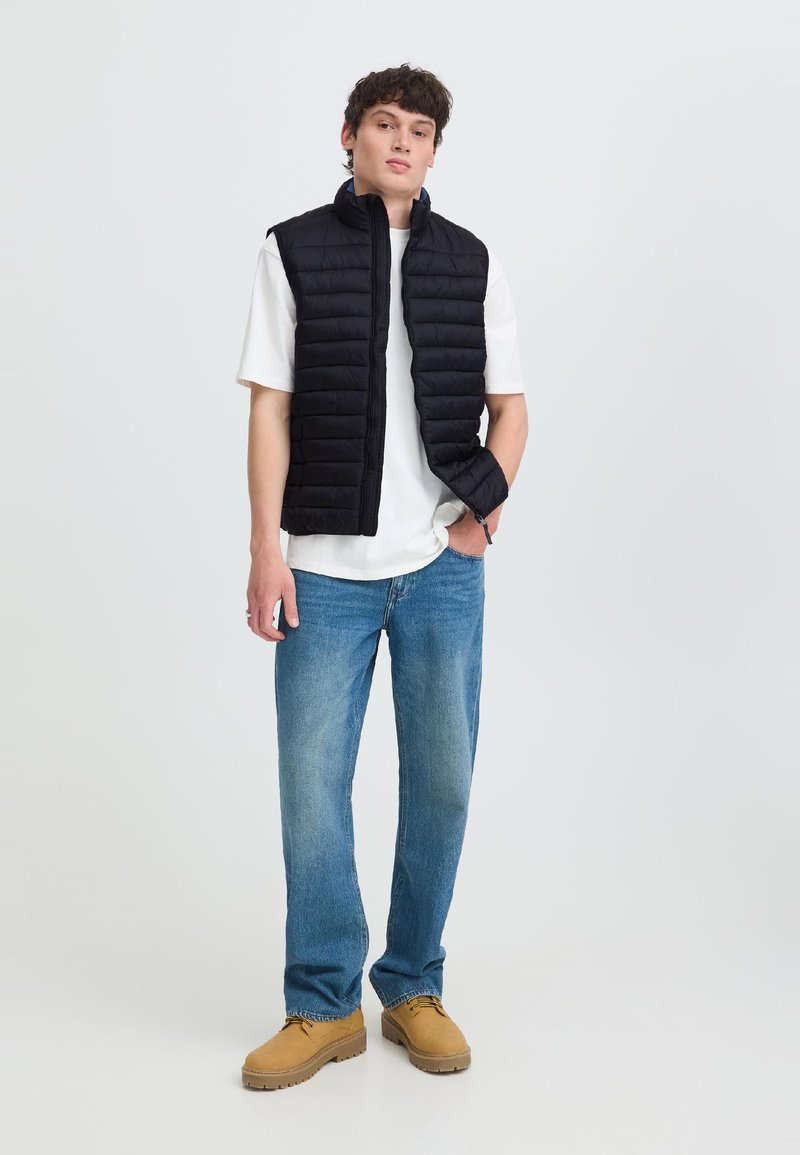Ung mand iført en sort quiltet vest over en hvid t-shirt, blå jeans og brune snørestøvler, stående med den ene hånd i lommen.