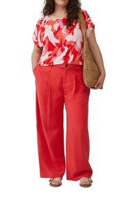 Frau trägt ein rotes und pinkes Oberteil mit abstraktem Muster, weite rote Hose mit Gürtel, beige Sandalen und hält eine braune geflochtene Schultertasche.