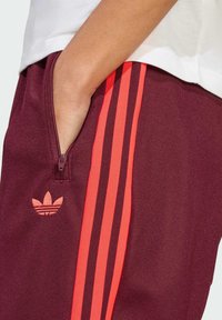 Bordeauxfarbene Jogginghosen mit leuchtend orangefarbenen Dreistreifen und gesticktem Logo. Sie verfügen über eine Reißverschlusstasche und einen strukturierten Stoff.