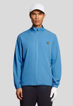 Mann trägt eine hellblaue Jacke mit Reißverschluss, weiße Kappe, schwarze Hose und einen weißen Handschuh an der rechten Hand, steht vor einem grauen Hintergrund.