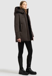 Giacca a vento marrone con cappuccio, dotata di zip frontale e tasche laterali. Abbinata a leggings neri e stivali neri al ginocchio.