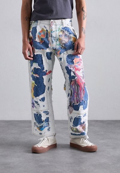 Jeans din denim uzat, cu petice colorate, având o croială lejeră și margini descusute. Asortate cu adidași bej cu tălpi maro.