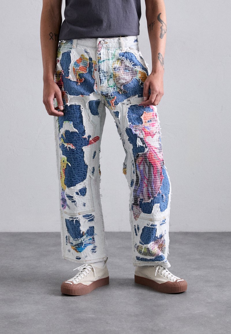 Distressed denim jeans with colorful patches, featuring a loose fit and frayed edges. Paired with beige sneakers with brown soles.