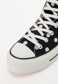 Svarta canvas sneakers med vita snören, prydda med stora rosa prickar och vit gummitå. Texturerad vit yttersula med ett mönstrat detaljer.